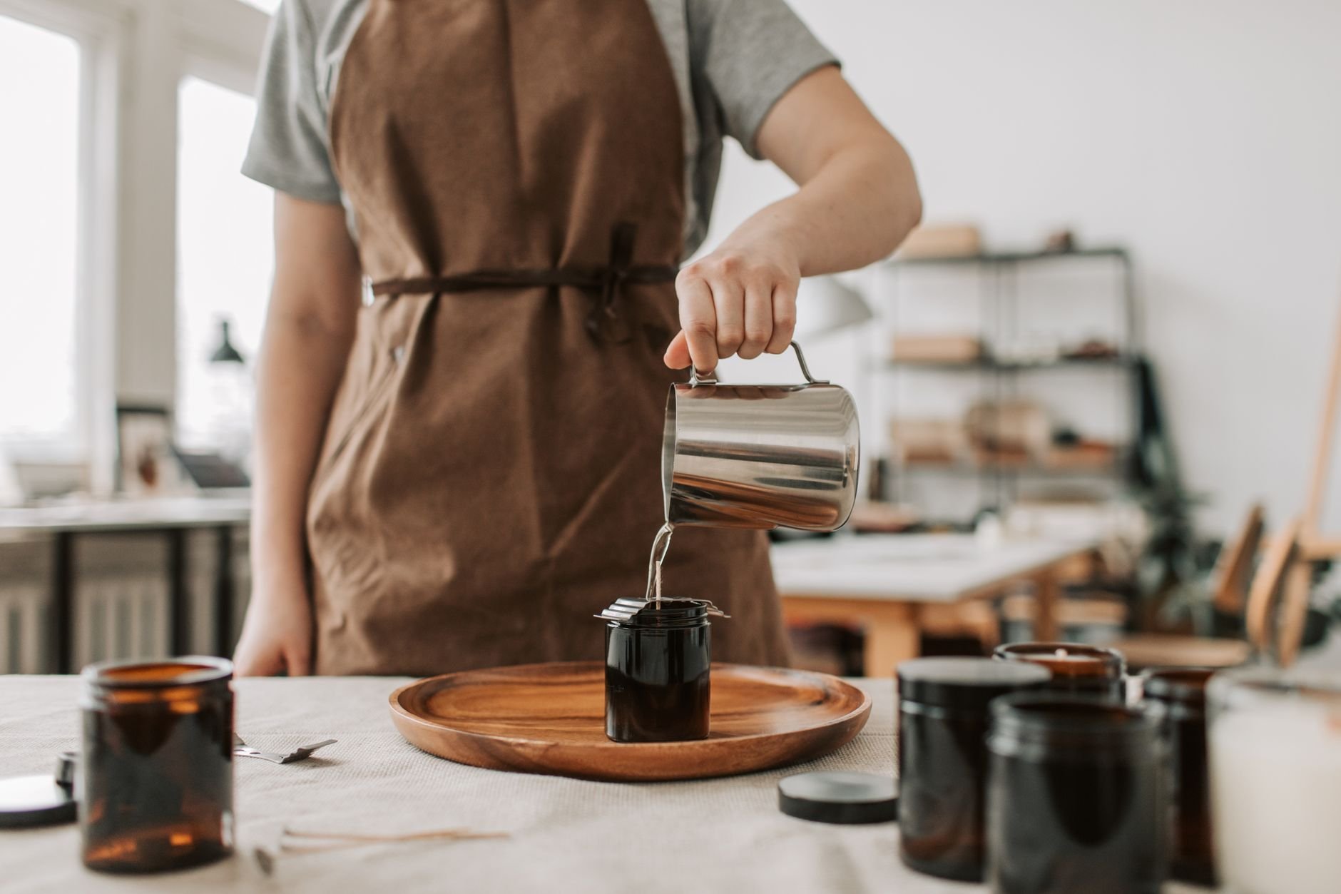 Person pouring wax into glass jars, creating handmade candles in a cosy workshop setting.