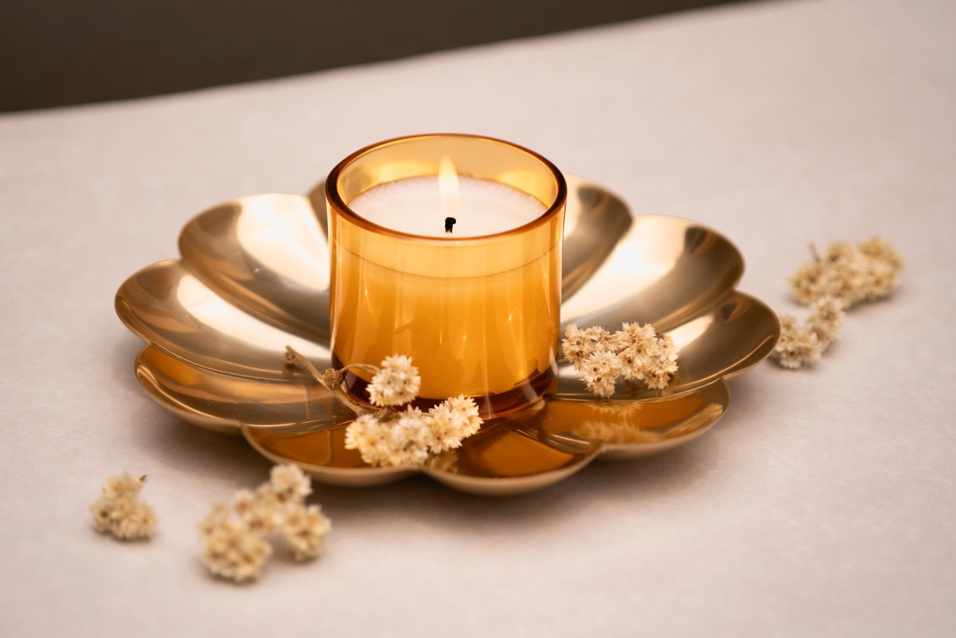 A lit candle in a golden holder surrounded by dried flowers on a white surface.
